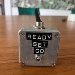 May include: A small, silver metal box with the words "READY SET GO" in black lettering. It has a black toggle switch on top and a small light. The box is secured with four screws.