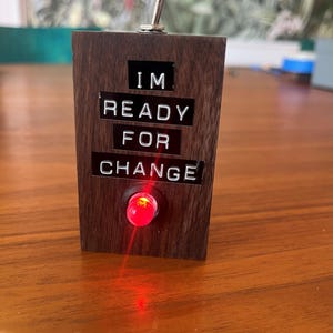 May include: A dark wooden block with the text "I'M READY FOR CHANGE" in white letters. A red light is illuminated at the bottom, and a silver toggle switch is at the top. The block is sitting on a wooden surface.