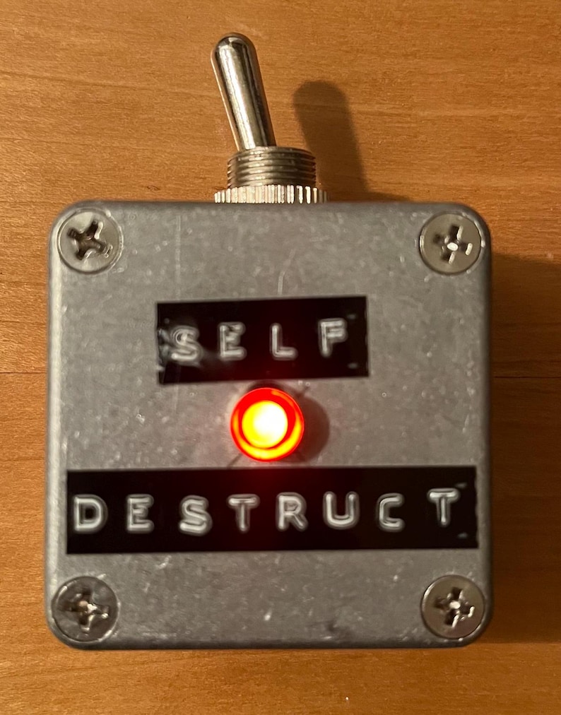 May include: A silver box with a red button and a toggle switch. The box has the words "SELF DESTRUCT" printed on it.