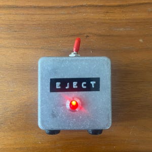 May include: A silver metal box with a red button and the word "EJECT" printed on it. The button is lit up red.