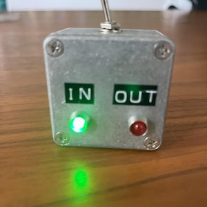 May include: A small, silver metal box with the words "IN" and "OUT" in green lettering. It has a toggle switch on top, a green light on the left, and a red light on the right. The box is secured with four screws.