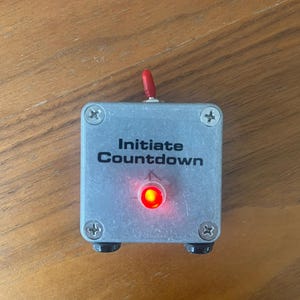 May include: A silver box with a red button and a red LED light. The box has the text "Initiate Countdown" printed on it.