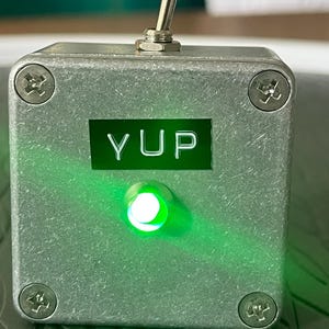 May include: A small, silver metal box with the word "YUP" illuminated in green. It features a toggle switch on top and a green light. The box is secured with four screws, and the overall design is industrial.