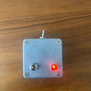 May include: A small, silver box with a red light on the right side and a knob on the left side. The box has a small, silver switch on the top.