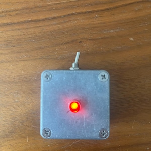 May include: A small grey metal box with a red LED light inside. The box has four screws on the sides and a small silver toggle switch on the top.