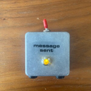 May include: A silver box with a red knob and a yellow light. The box has the text "message sent" printed on it.