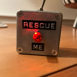 May include: A small, silver metal box with the words "RESCUE ME" in black lettering. A red light is illuminated below the word "RESCUE". A silver toggle switch is on top. The box is secured with four screws.