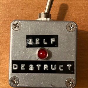 May include: A silver box with a red light and a toggle switch. The box has the words "SELF DESTRUCT" printed on it.