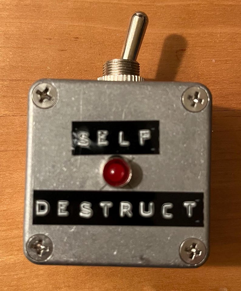 May include: A silver box with a red light and a toggle switch. The box has the words "SELF DESTRUCT" printed on it.