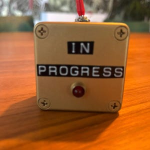 May include: A gold-coloured box with the words "IN PROGRESS" displayed. It features a red button and a red toggle switch. Four screws secure the face. The box is on a wooden surface.