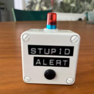 May include: A white square plastic box with four corner screws, featuring a black label with the white text STUPID ALERT. A small black circular button is positioned below the text, and a red translucent light sits on top with a blue base.