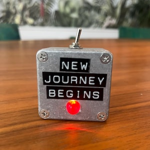 May include: A small, silver metal box with the words "NEW JOURNEY BEGINS" in white lettering. It has a toggle switch on top and a red light illuminated on the front. The box is secured with four screws.