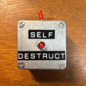 May include: A square silver metal box featuring a red toggle switch on top and a red indicator light in the center. The front panel displays the text SELF in white letters on a black background above the light and DESTRUCT below the light.