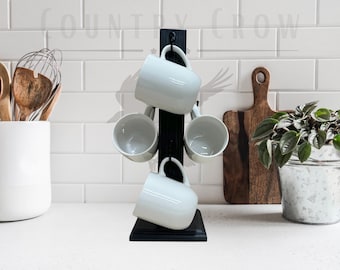 May include: A black wooden mug rack with four white ceramic mugs hanging from hooks. The rack is standing on a white countertop.