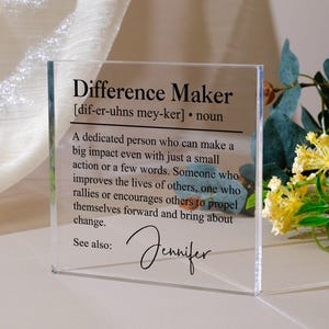 Puede incluir: Un bloque acrílico transparente con la inscripción "Difference Maker" y su definición en negro. El bloque está firmado "Jennifer". El fondo incluye flores y una cortina blanca translúcida.