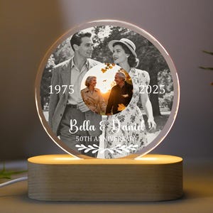 Può includere: Un espositore fotografico rotondo e illuminato con un'immagine in bianco e nero di una coppia, con gli anni 1975 e 2025. Include una foto a colori più piccola di una coppia più anziana. La base è in legno chiaro e il testo recita "Bella & Daniel 50th Anniversary."