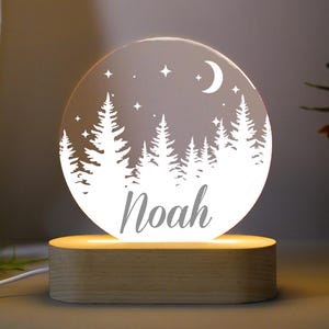 May include: A round, illuminated night light with a forest scene and the name "Noah" etched into the surface. The design features trees, stars, and a crescent moon. The light sits on a rectangular wooden base.