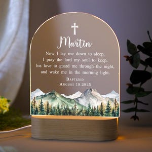 May include: A personalised night light with a wooden base and an arched acrylic panel. The panel features a cross, the name "Martin", a prayer, and the date "August 19, 2025". A mountain and forest scene is at the bottom.