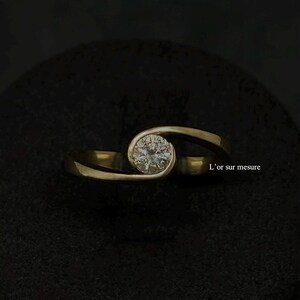 Puede incluir: Un anillo de oro con un solo diamante redondo engastado en el centro. La banda es un diseño simple y curvo. El anillo está sobre un fondo negro. L'or sur mesure.