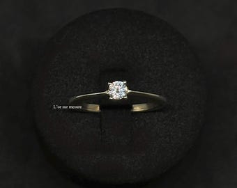 Könnte beinhalten: Ein goldener Solitär-Verlobungsring mit einem runden Diamanten. Der Ring ist auf einem dünnen Band gefasst und wird auf einer schwarzen Oberfläche präsentiert. Der Text "L'or sur mesure" ist sichtbar.