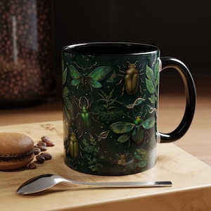 Peut inclure: Une tasse en céramique noire avec un motif vert et or représentant un motif répété de scarabées et de feuilles. La tasse a une anse noire.