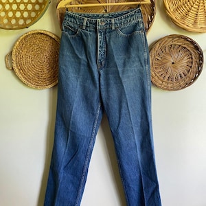 May include: Blue denim jeans hanging on a wooden hanger. The jeans have a classic five-pocket design and a button and zipper closure. The denim is a medium wash with slight fading.