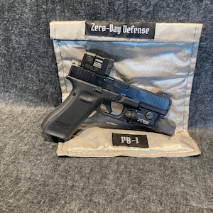 May include: A black handgun with a red dot sight and a flashlight attached. The gun is resting on a gray fabric bag with the text "Zero-Day Defense" and "PB-1" printed on it.