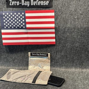 May include: A silver pouch with a black zipper and the text "Zero-Day Defense" on the front. The pouch is open and a black phone is partially inside. An American flag is in the background.