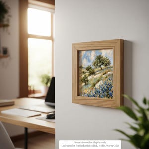 a framed painting of a landscape with trees and flowers hanging on a wall in a room with a desk and a window.