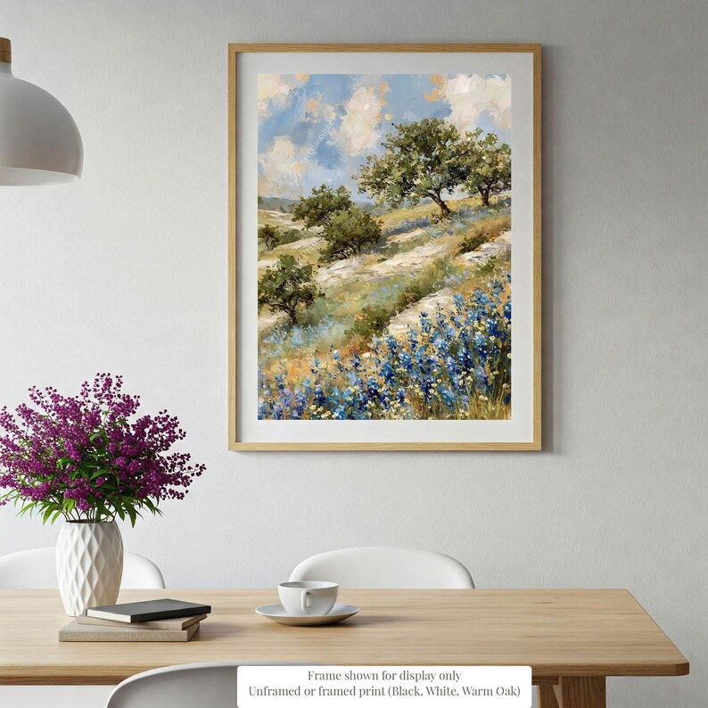 a framed painting of a scenic landscape with trees, flowers, and a blue sky with clouds hanging on a wall above a wooden table with a vase of purple flowers and a stack of books.