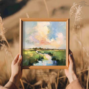 A person's hands holding a framed painting of a landscape with a river and green fields, against a blurred natural background.