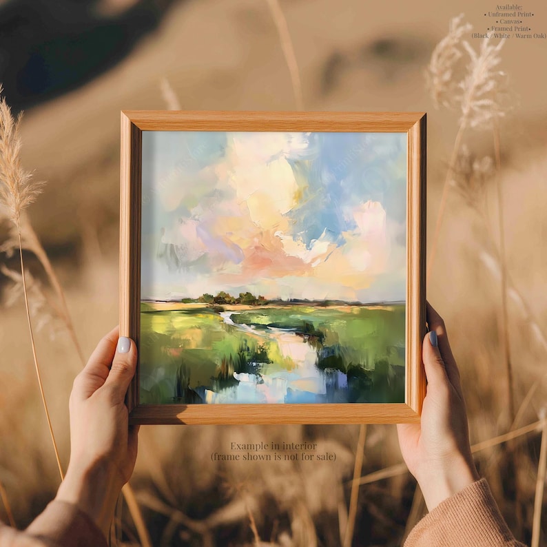 A person's hands holding a framed painting of a landscape with a river and green fields, against a blurred natural background.