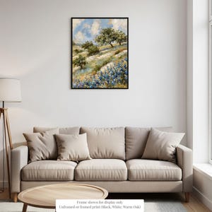 a living room with a beige sofa and a wooden coffee table. Above the sofa, there is a framed painting depicting a landscape with trees and flowers. The painting is hanging on a white wall, and the frame is described as "Frame shown for display only" and "Unframed or framed print (black, white, warm oak)."