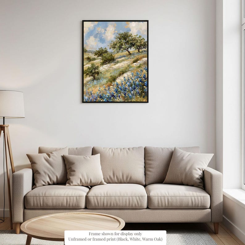 a living room with a beige sofa and a wooden coffee table. Above the sofa, there is a framed painting depicting a landscape with trees and flowers. The painting is hanging on a white wall, and the frame is described as "Frame shown for display only" and "Unframed or framed print (black, white, warm oak)."