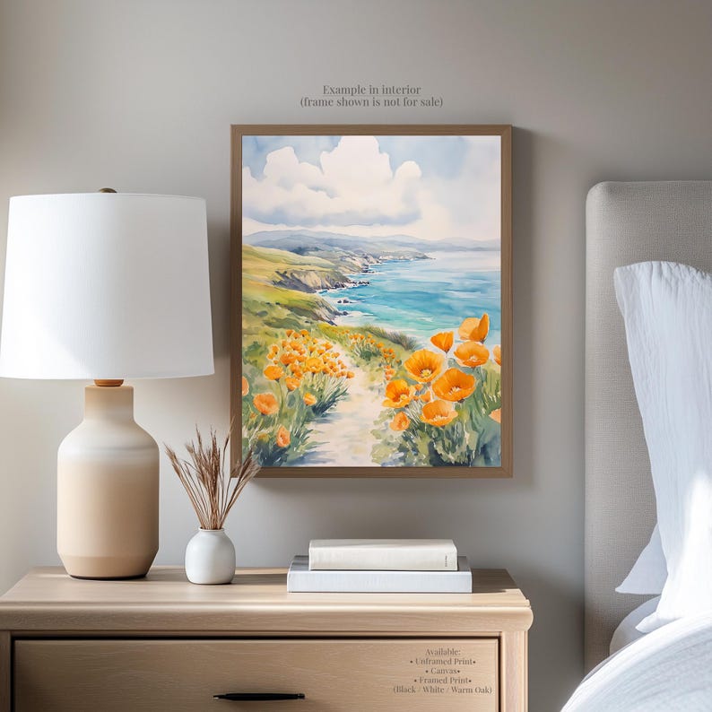 a bedroom setting with a framed painting of a coastal landscape hanging on the wall above a wooden nightstand. The painting depicts a path leading through a field of orange flowers, with a body of water visible in the background. The nightstand has a lamp, a vase with dried flowers, and a stack of books.