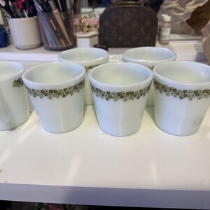 以下が含まれることがあります： 緑色の花柄が縁に施された白いセラミック製のマグカップが6つ。マグカップは白い表面に一列に並んでいます。