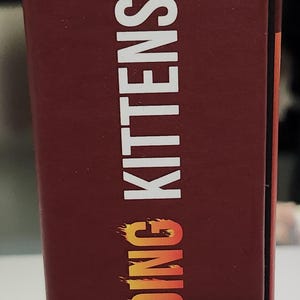 Op de afbeelding: Een kastanjebruine doos met de tekst "EXPLODING KITTENS" in wit met een oranje verloop. Bovenop staat een cartoon kat silhouet. De doos is voor een kaartspel.