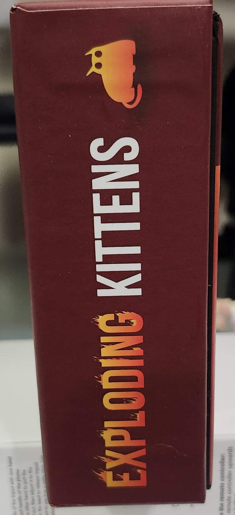 Op de afbeelding: Een kastanjebruine doos met de tekst "EXPLODING KITTENS" in wit met een oranje verloop. Bovenop staat een cartoon kat silhouet. De doos is voor een kaartspel.