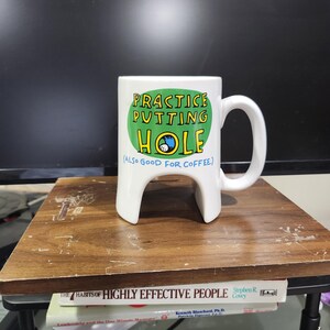 May include: White ceramic mug with a unique design, featuring a handle and a base that resembles a golf tee. The mug has the text "PRACTICE PUTTING HOLE (ALSO GOOD FOR COFFEE)" in green, blue, and yellow.