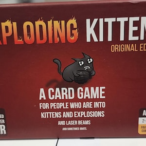May include: A red box for the Exploding Kittens card game. The title is in orange and white, with a cartoon cat illustration. Text includes "A Card Game" and "For People Who Are Into Kittens and Explosions".
