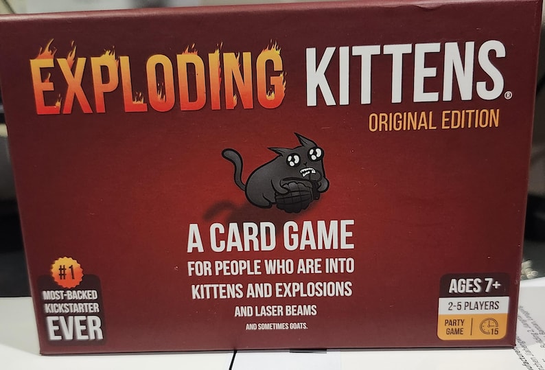 Op de afbeelding: Rode doos voor het Exploding Kittens kaartspel. De titel is in oranje en wit, met een cartoon kat illustratie. Tekst bevat "A Card Game" en "For People Who Are Into Kittens and Explosions".