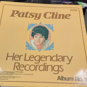 May include: A vintage album cover featuring Patsy Cline. The cover is a light tan color with the title "Patsy Cline" in brown lettering. A portrait of Cline is in a blue and white ornate frame. The text "Her Legendary Recordings" is below the portrait.