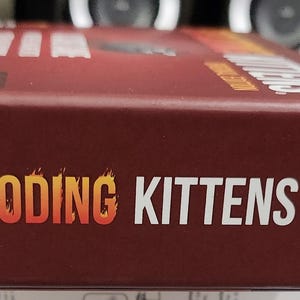Op de afbeelding: Een kastanjebruine speldoos met de woorden "EXPLODING KITTENS" in oranje en wit. Het woord "EXPLODING" heeft een vlamontwerp. Een cartoonkat staat aan de rechterkant van de doos. De speldoos staat op een wit oppervlak.