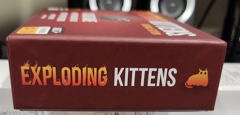 Op de afbeelding: Een kastanjebruine speldoos met de woorden "EXPLODING KITTENS" in oranje en wit. Het woord "EXPLODING" heeft een vlamontwerp. Een cartoonkat staat aan de rechterkant van de doos. De speldoos staat op een wit oppervlak.
