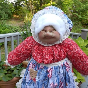 May include: A handmade doll dressed in a red and white patterned top and a blue denim-style skirt adorned with American flags. The doll has a white bonnet, glasses, and a smiling face. A wooden tag is attached to the skirt.