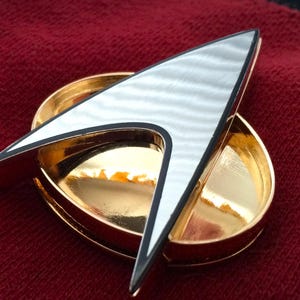 Op de afbeelding: Een goud-zilveren Starfleet-insigne. Het heeft een zilveren pijlpuntontwerp met zwarte rand, geplaatst op een gouden ovale basis. De insigne wordt gepresenteerd op een rode stoffen achtergrond, die de metallic glans en details benadrukt.