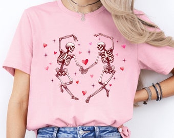 May include: Pink t-shirt with a graphic of two skeletons dancing and holding hands. The skeletons are surrounded by small pink hearts and stars.