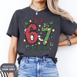 May include: Heather dark grey t-shirt with a festive design. The graphic features the numbers "67" in red and green, adorned with Christmas lights and surrounded by stars, gift boxes, and the words "Six Seven".