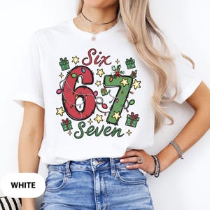 May include: White t-shirt with a festive design featuring the numbers "6" and "7" decorated with Christmas lights. The word "Six" is above the "6" and "Seven" is below the "7". The design includes gift boxes and stars.