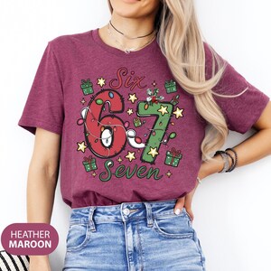 May include: Heather maroon t-shirt with the text "Six 6 Seven" in a festive design. The numbers are decorated with Christmas lights and surrounded by gift boxes and stars. A fun birthday shirt.